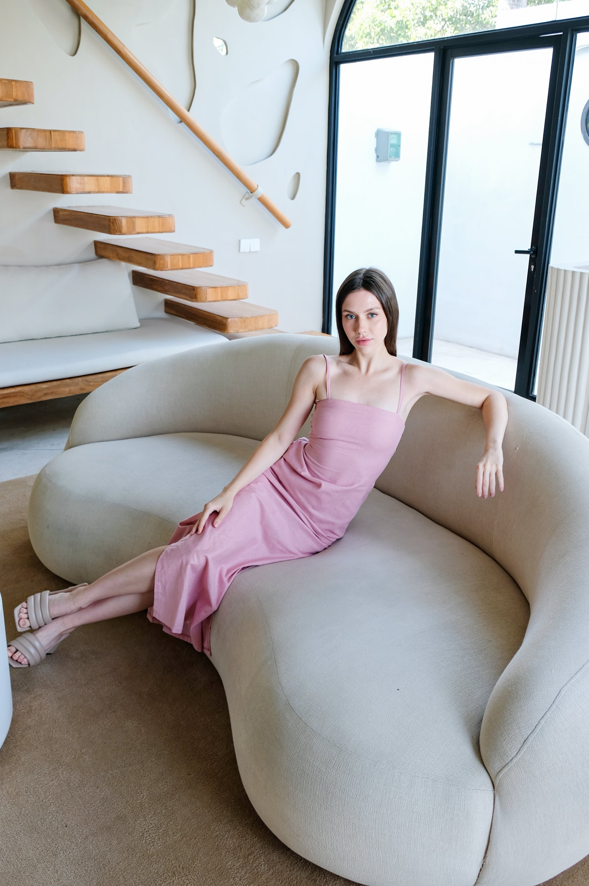 Model wearing pink linen dress by Una Linen Havn is sitting comfortably on white sofa.