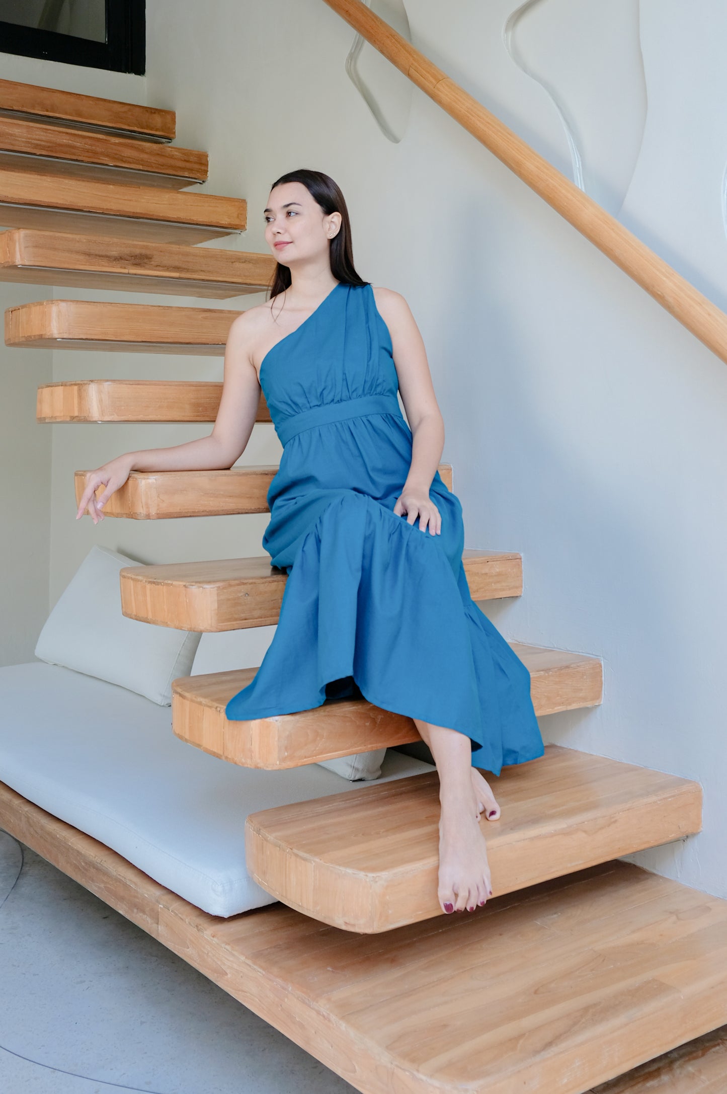 Blue variant of one-shoulder dress displayed by a model sitting on wooden staircase.